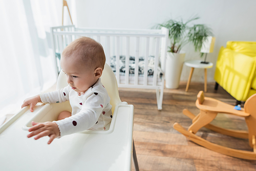 toddler boy sitting in baby chair near blurred crib and rocking horse