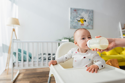 woman with milk in baby bottle feeding son near blurred crib