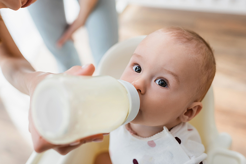 high angle view of toddler boy drinking milk from baby bottle near blurred mom
