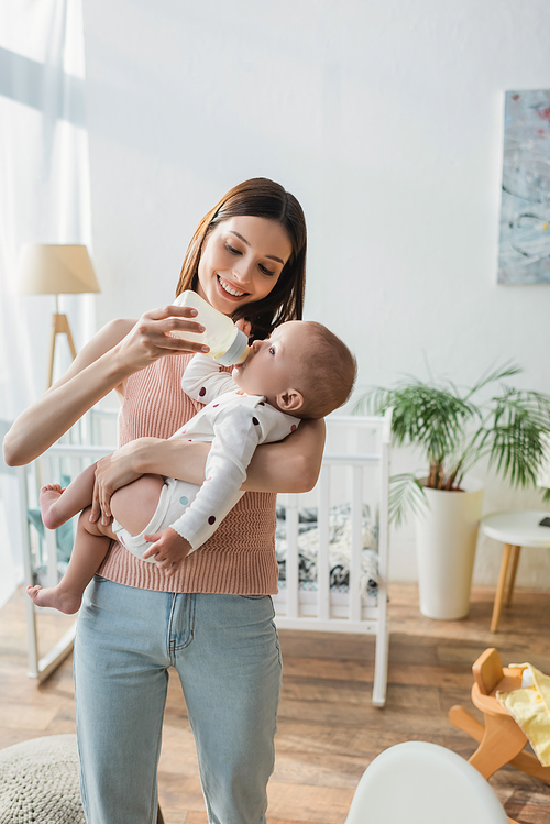 happy woman feeding little son with milk while standing near blurred crib
