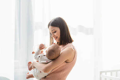 happy brunette woman feeding toddler from baby bottle while standing near white curtain