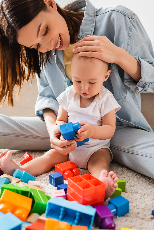 smiling woman touching head of toddler son while playing with building blocks on floor