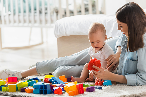 woman holding building block while playing with baby boy on floor in bedroom