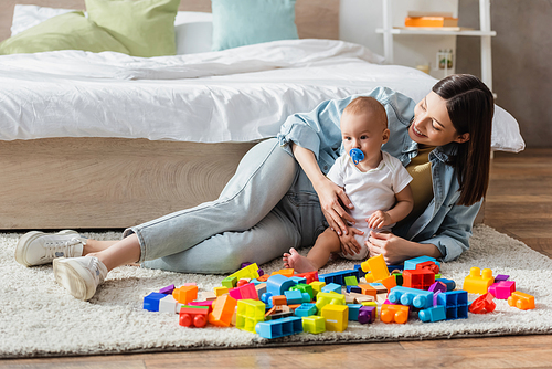 baby boy with pacifier and his happy mom playing with colorful building blocks on floor