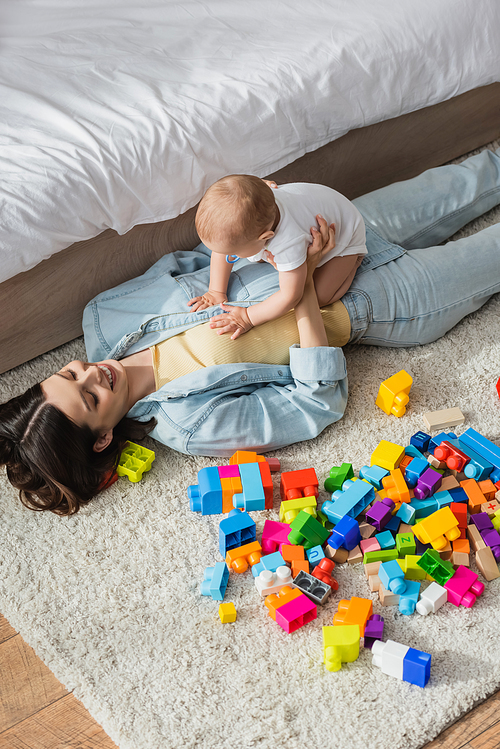 high angle view of happy woman lying on floor near building blocks while playing with toddler son