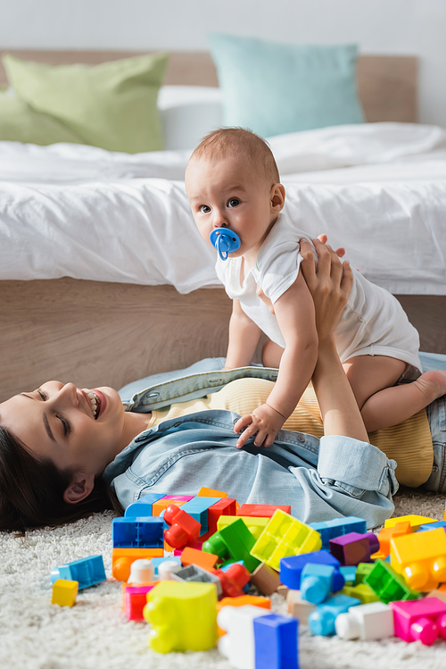 happy woman playing with toddler son near building blocks on floor at home