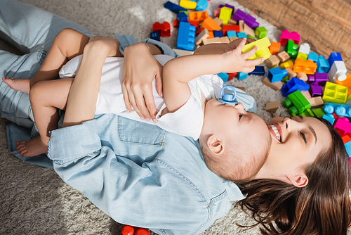 top view of cheerful woman with closed eyes embracing little son near building blocks on floor
