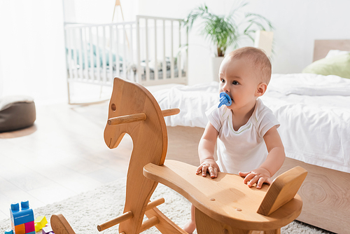 toddler child with pacifier standing near rocking horse, blurred bed and crib