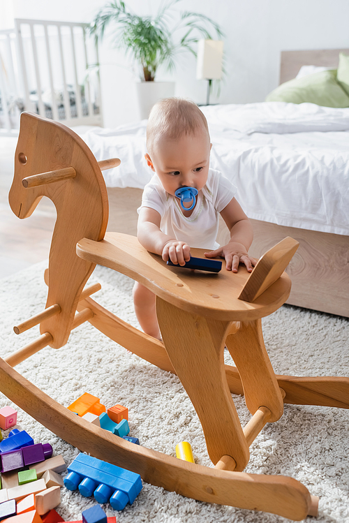 toddler kid with pacifier standing near rocking horse and building blocks on floor