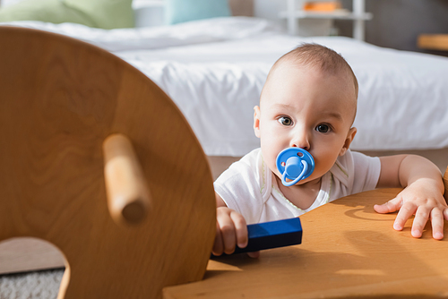 baby with pacifier holding toy building block while  near blurred rocking horse
