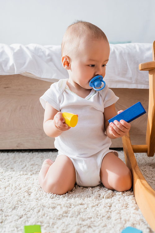 little kid with pacifier holding toy building blocks while sitting on floor carpet