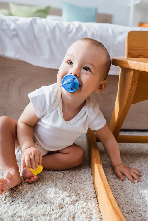 cheerful little boy with pacifier holding toy building block while sitting on floor
