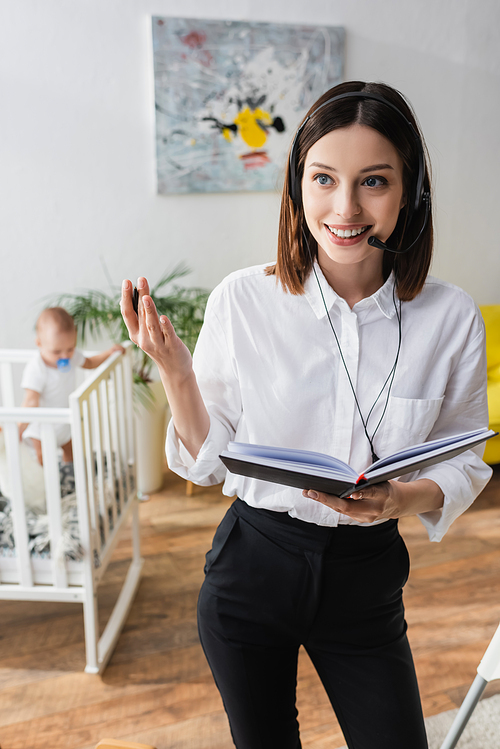 happy woman in headset holding notebook while working near little son in crib on blurred background