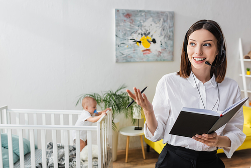 smiling woman talking in headset near baby boy in crib on blurred background