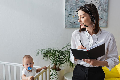 woman in headset writing in notebook while working at home near baby in crib