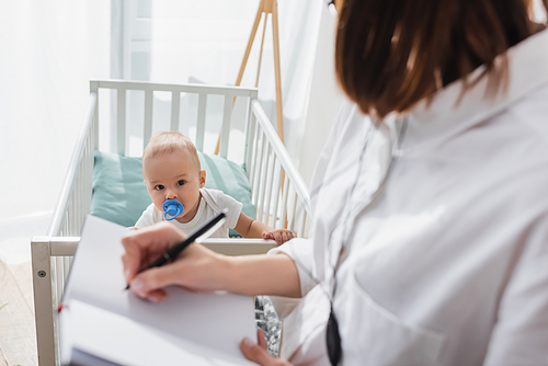 blurred woman writing in notebook while working near child in crib