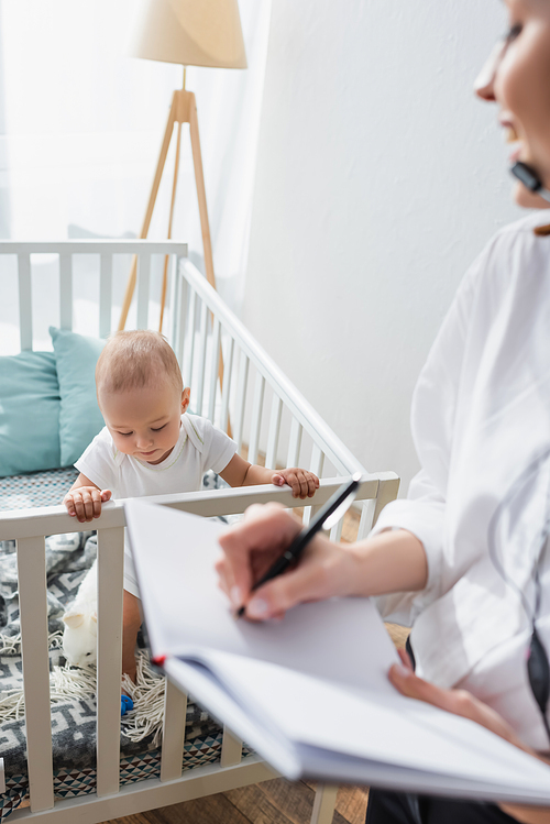 blurred woman writing in notebook near toddler kid in baby crib