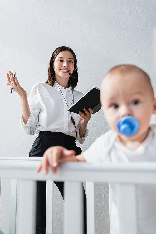 happy woman with notebook working in headset near blurred toddler kid in crib