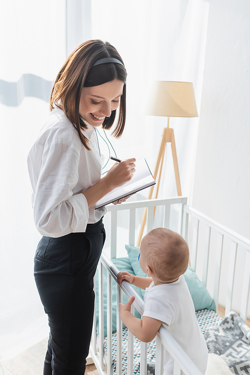 cheerful woman in headset holding pen and notebook while standing near little son in crib