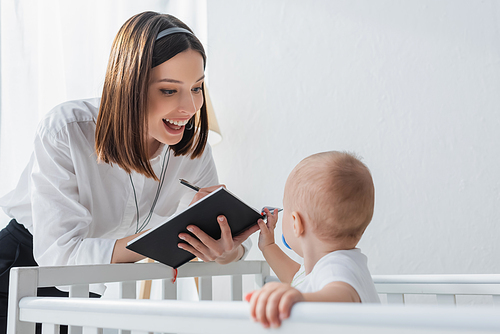 happy woman in headset writing in notebook near toddler son in crib