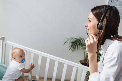 happy woman talking in headset near blurred toddler child in crib