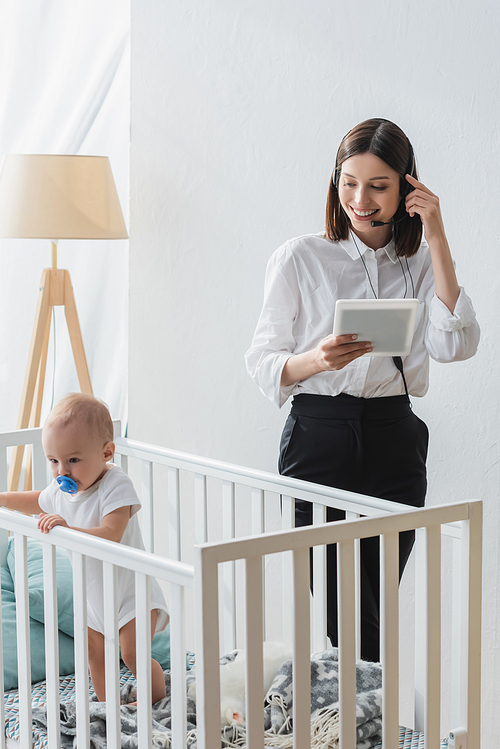 smiling woman with digital tablet working in headset near toddler child in crib