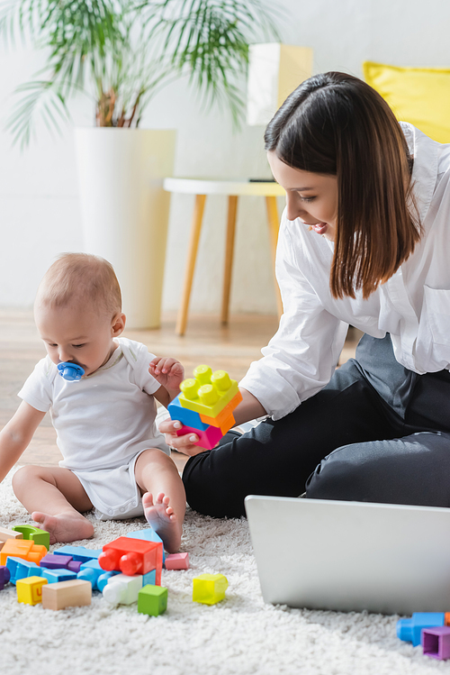 brunette woman holding building block while sitting on floor near little son and laptop