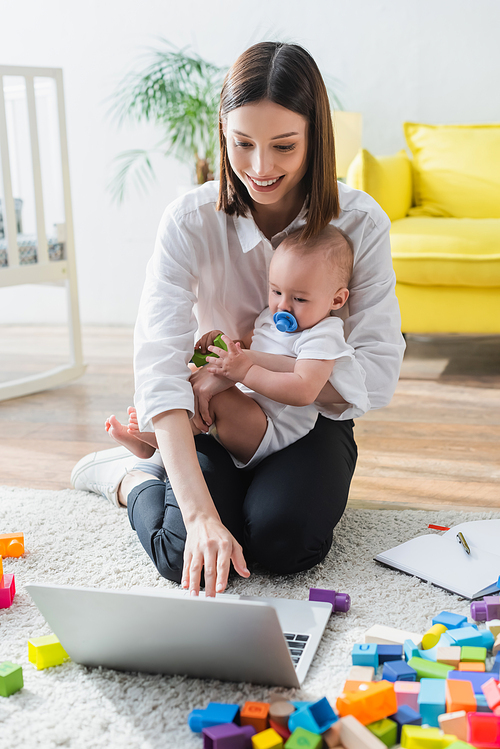 smiling woman using laptop while sitting on floor with son near building blocks