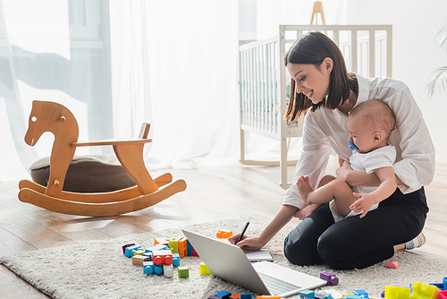 smiling woman writing in notebook while sitting on floor with son near laptop and building blocks