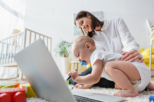 toddler boy holding pen near happy mom and laptop on floor