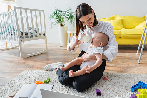 happy woman sitting on floor near laptop and feeding little son from baby bottle