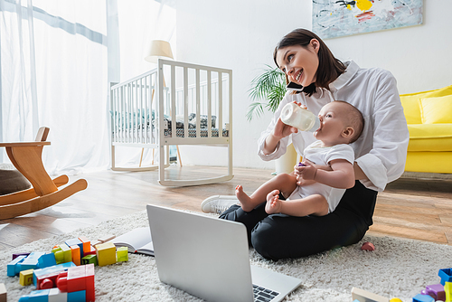 smiling woman talking on smartphone while feeding son on floor near laptop and building blocks