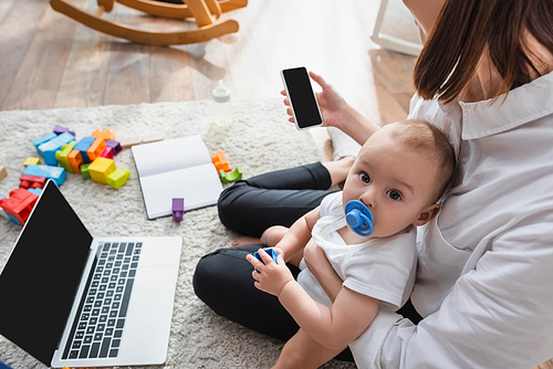 woman with smartphone sitting on floor with baby boy near laptop and building blocks