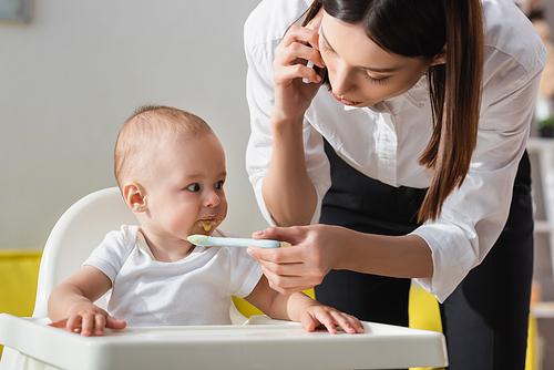 woman talking on mobile phone while feeding toddler son with puree