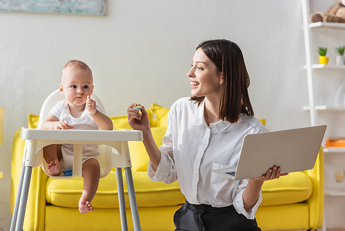 toddler boy in baby chair showing thumb up near happy mom with spoon and laptop