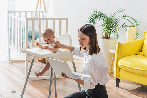 brunette woman sitting on floor with laptop while feeding son in baby chair