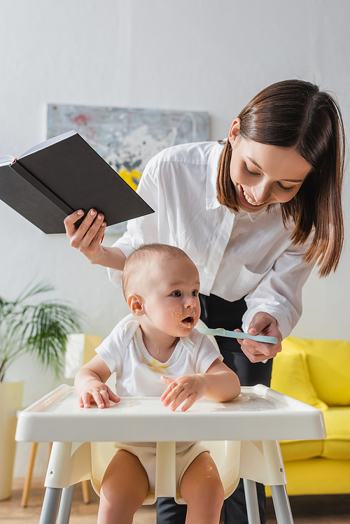 cheerful woman with notebook feeding little son with puree at home