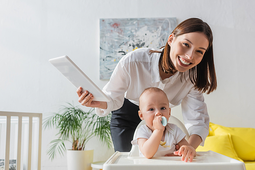 cheerful woman with digital tablet  near little son sitting in baby chair with spoon