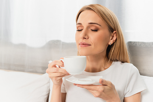 pleased blonde woman holding cup of coffee in bedroom