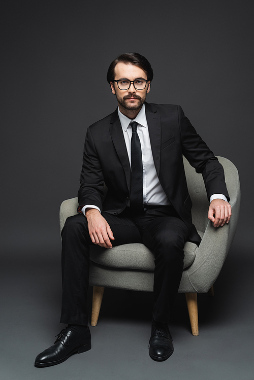 businessman in suit and glasses sitting on armchair on dark grey