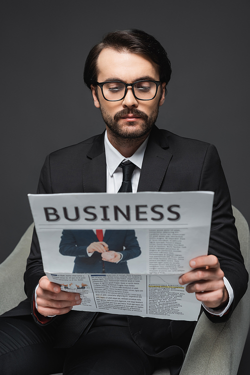 businessman in suit and glasses sitting on armchair and reading business newspaper on dark grey