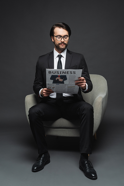 full length of businessman in suit and glasses sitting on armchair and reading business newspaper on dark grey