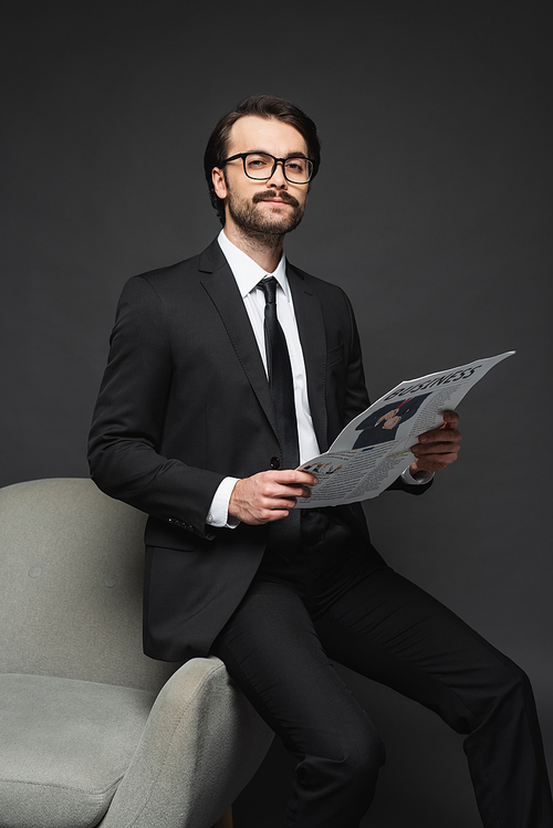 businessman in suit and glasses leaning on armchair and holding business newspaper on dark grey