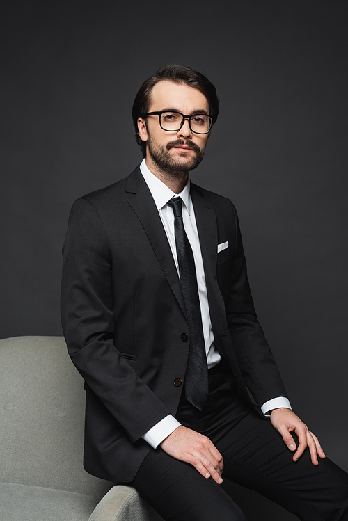 businessman in formal wear and glasses leaning on armchair on dark grey
