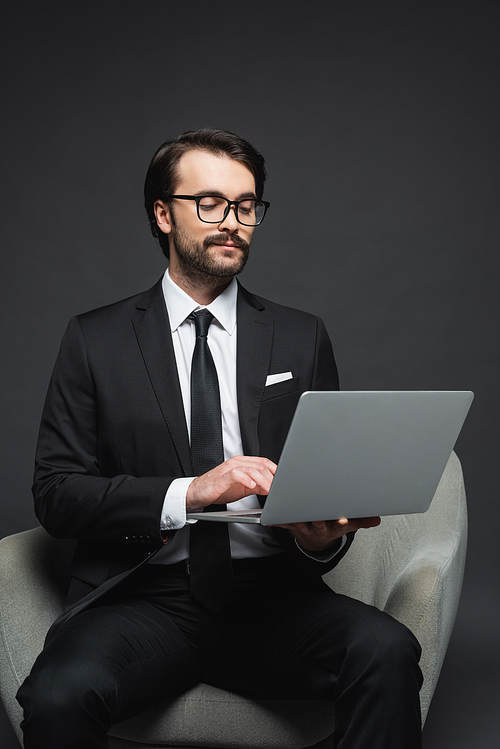 businessman in suit and glasses sitting on armchair and using laptop on dark grey