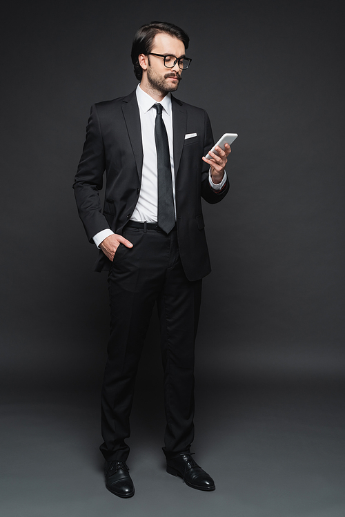 full length of businessman in suit and glasses standing with hand in pocket and using smartphone on dark grey