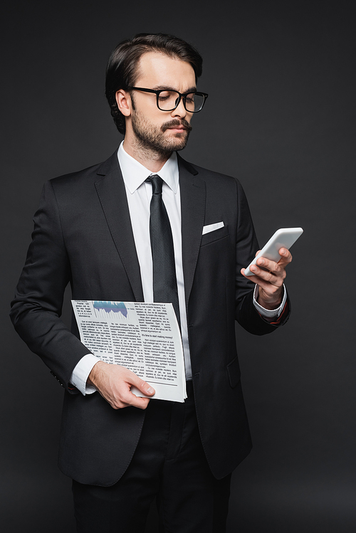 businessman in suit and glasses holding newspaper and using smartphone on dark grey