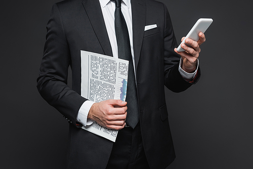 cropped view of businessman in suit holding newspaper and using smartphone on dark grey