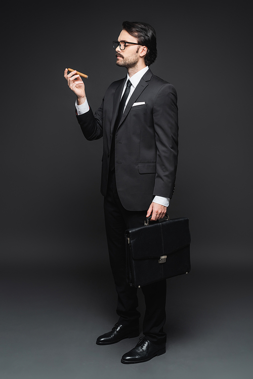 full length of man in suit and glasses holding cigar and leather briefcase on dark grey
