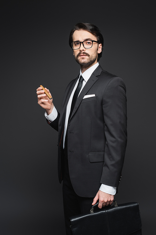 businessman in suit and glasses holding cigar and leather briefcase on dark grey